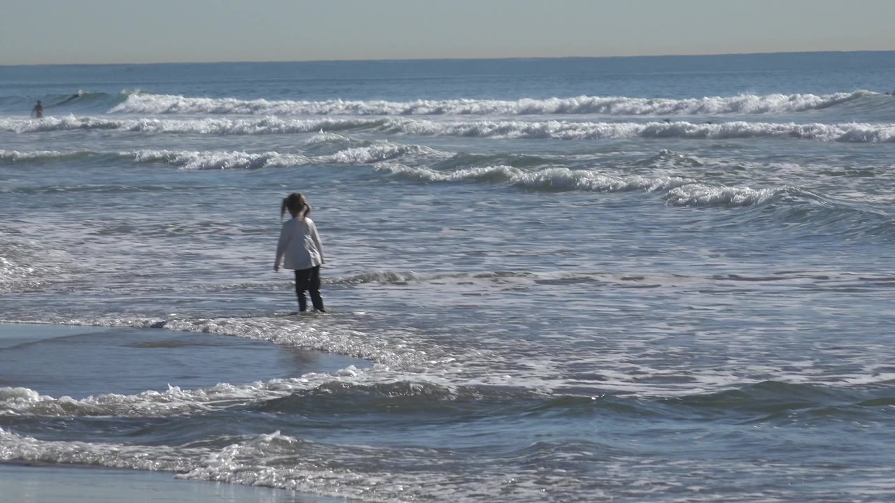 At Huntington Beach, California, the girl stands farther right in the shallow surf as a low wave line curves across the foreground.