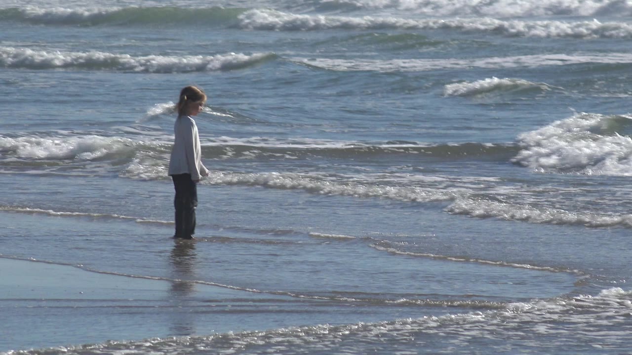 A young girl in a white shirt stands near the left side of the Huntington Beach shoreline as small waves roll in behind her.
