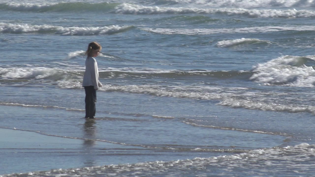Girl facing the ocean as a foamy wave breaks behind her.