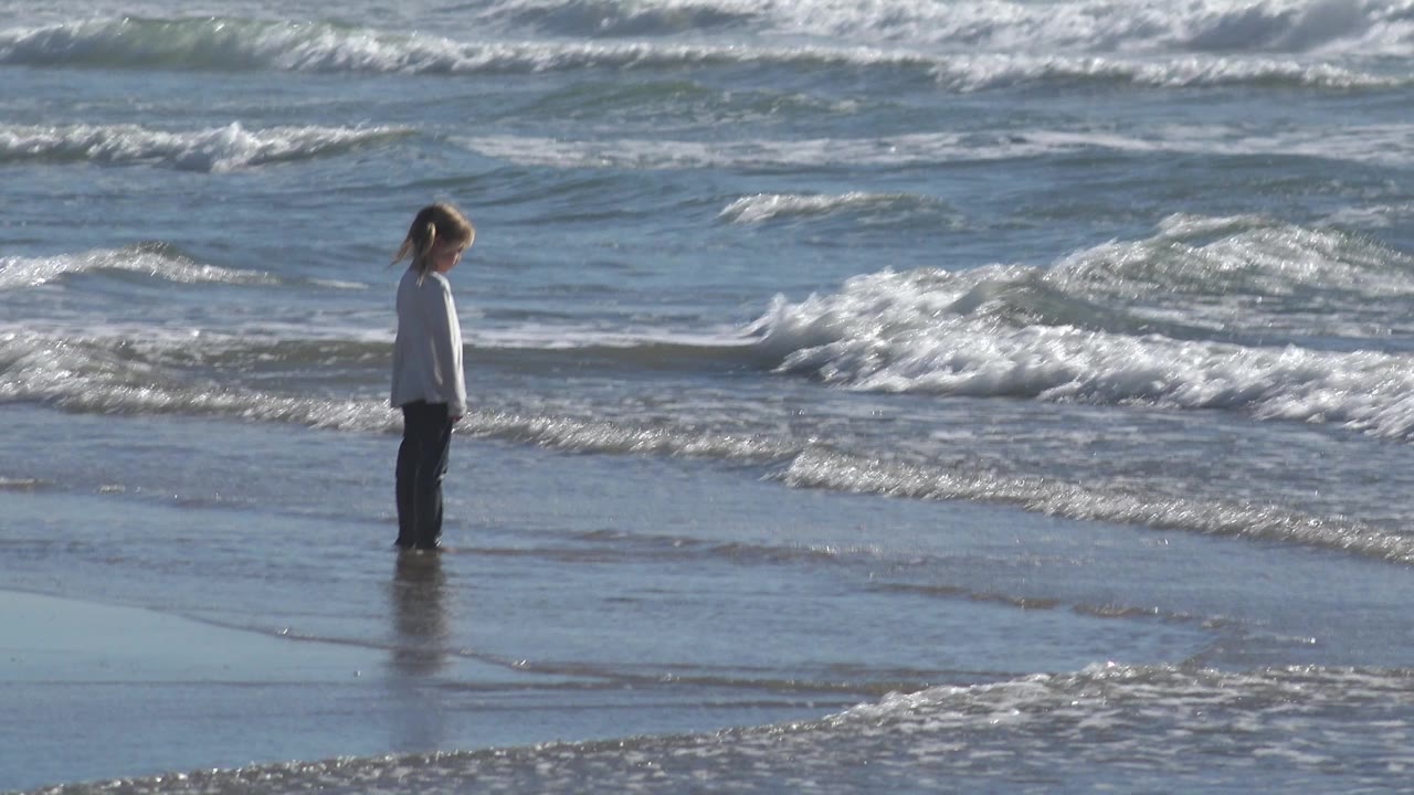 A young girl stands near the right side of the Huntington Beach frame, with choppy blue-gray waves and white foam behind her.