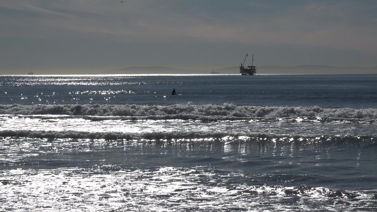 Huntington Beach waterline with bright highlights across the midground, low foamy waves in front, a far platform-like structure, and no visible people.