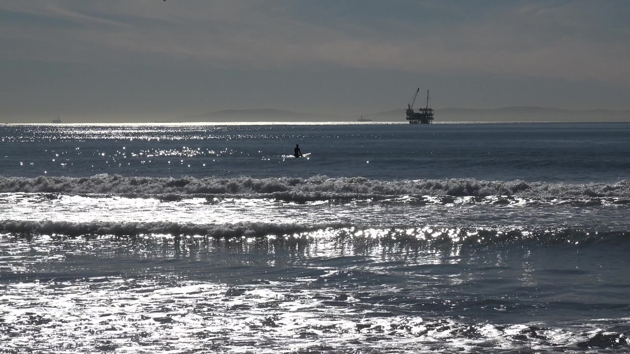 Huntington Beach surf frame with a broad bright reflection on the water, a breaking wave across the foreground, distant offshore structure, and no visible people.