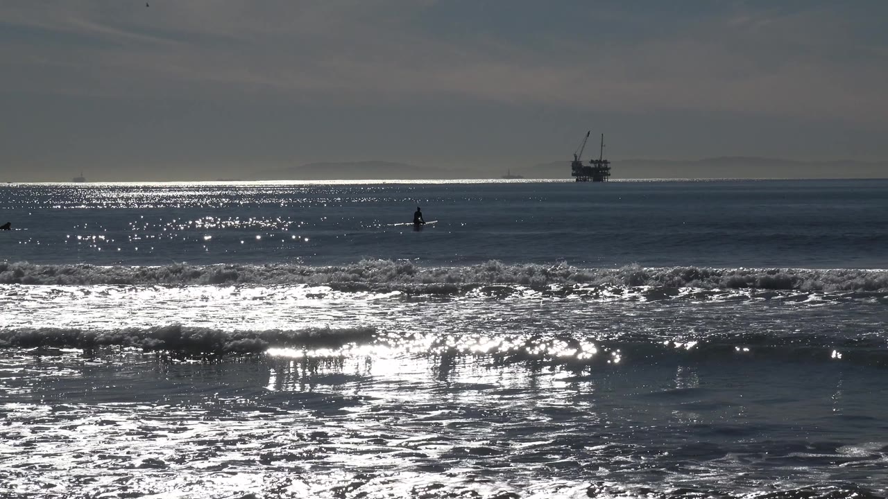 Huntington Beach ocean at 0:03 with reflective water, a low foreground wave, a distant platform-like structure near the horizon, and no visible people.