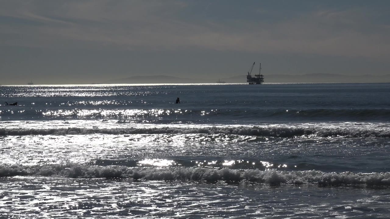 Huntington Beach ocean view with bright sun glare across the water, a low breaking wave near shore, an offshore platform on the horizon, and no visible people.