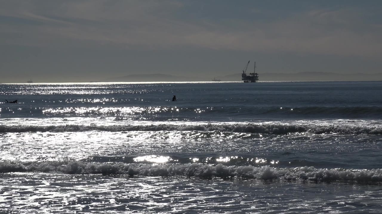Huntington Beach surf under strong reflected light, with foam forming in the foreground, the offshore platform centered near the horizon, and no visible people.