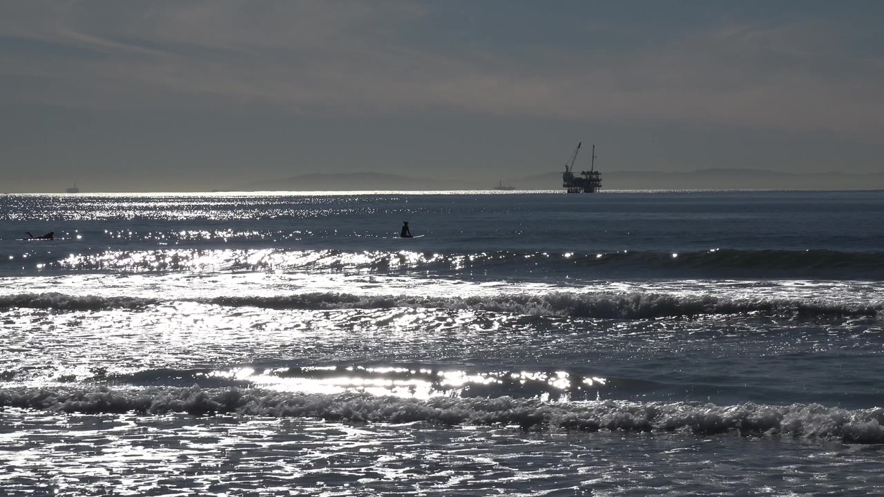 Huntington Beach water fills the frame with silver highlights, a darker swell crossing the midground, an offshore platform at the horizon, and no visible people.