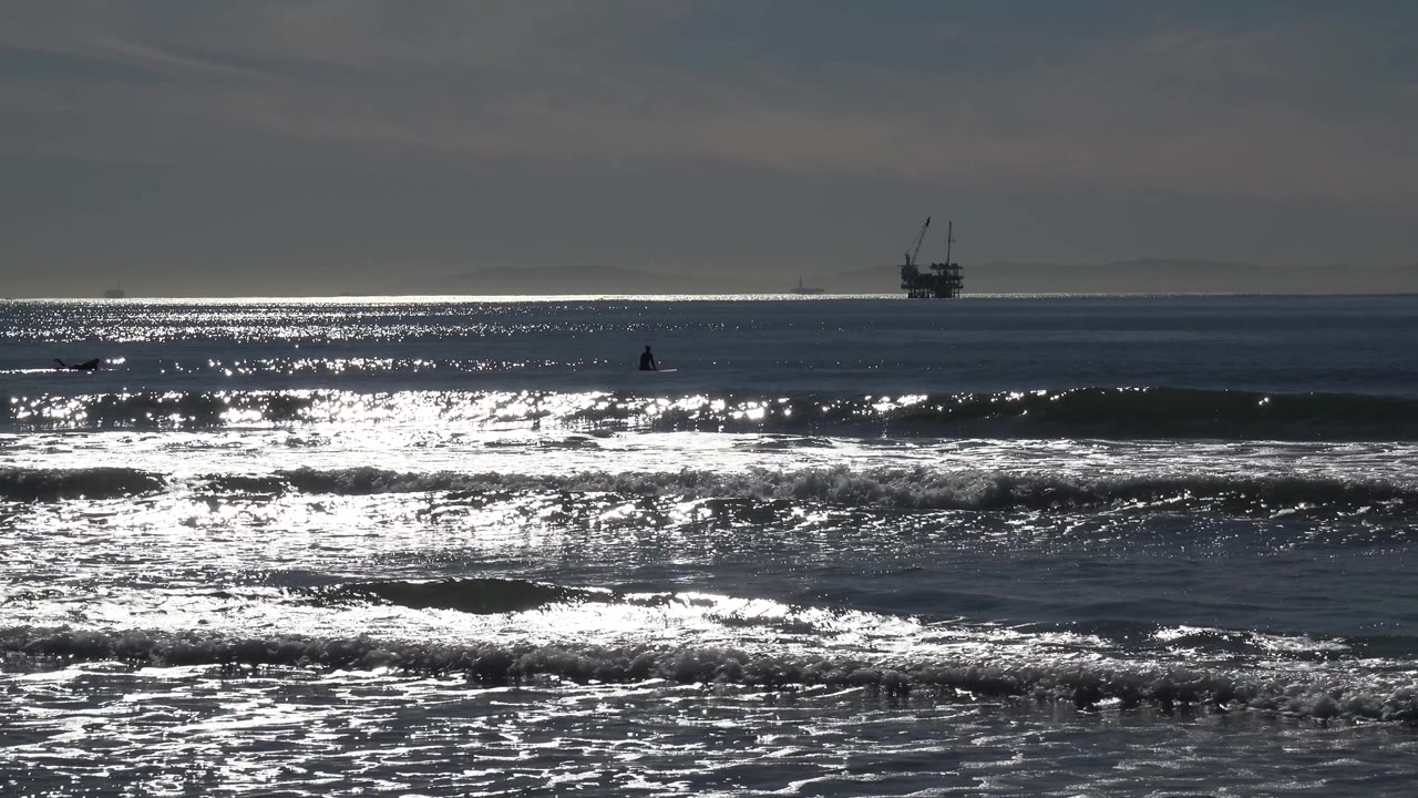 Shorebreak foam and sunlit ocean with a platform offshore.