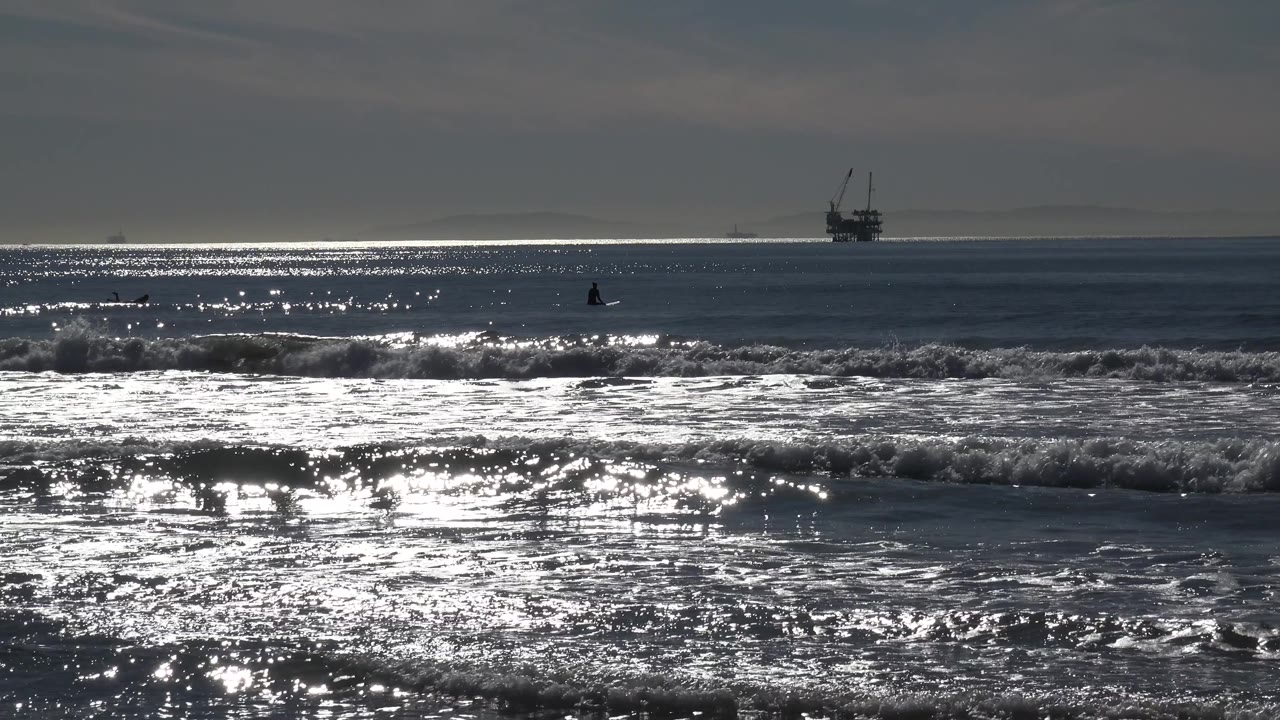 At Huntington Beach, foamy waves spread across the foreground as bright reflected light runs across the midground water, with the distant offshore structure still visible.