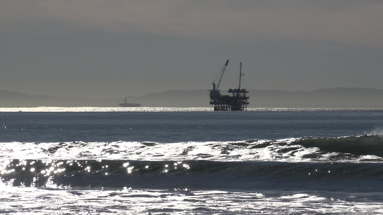 Huntington Beach surf breaks in bright backlight with a dark offshore industrial platform near the horizon and no visible people.