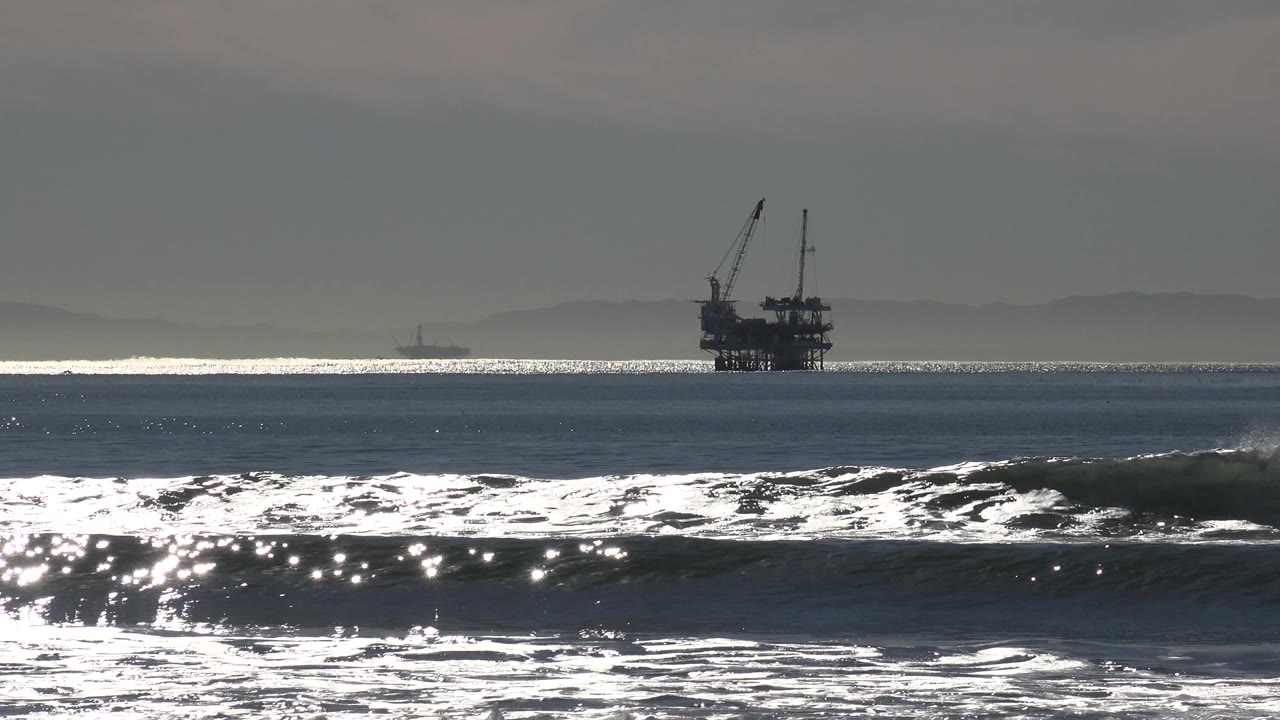 A low wave line crosses the Huntington Beach foreground while sunlit water bands and a silhouetted platform sit farther offshore.