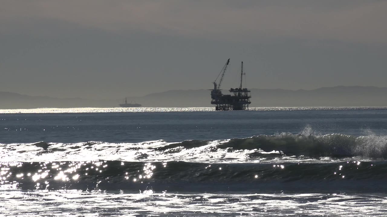 White surf flashes in the foreground as the offshore platform remains a dark silhouette against the reflective Huntington Beach water.