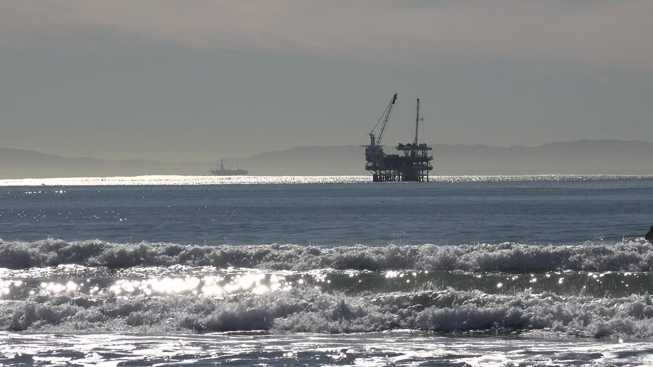Offshore oil rig silhouetted beyond bright breaking surf at Huntington Beach, California, with no visible people in this opening frame.