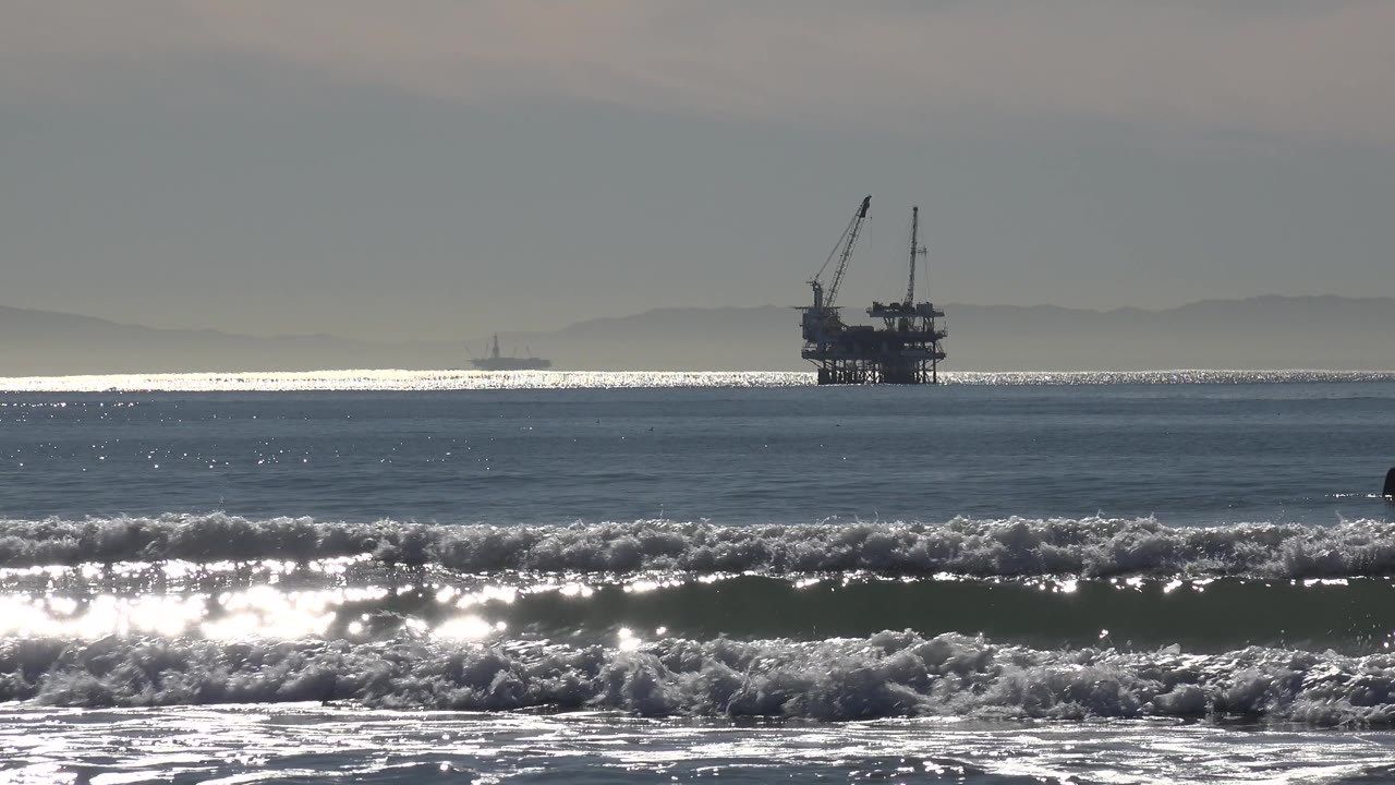 The offshore oil rig remains in the distance at Huntington Beach, California, while a small dark surfer or swimmer appears near the breaking wave line.
