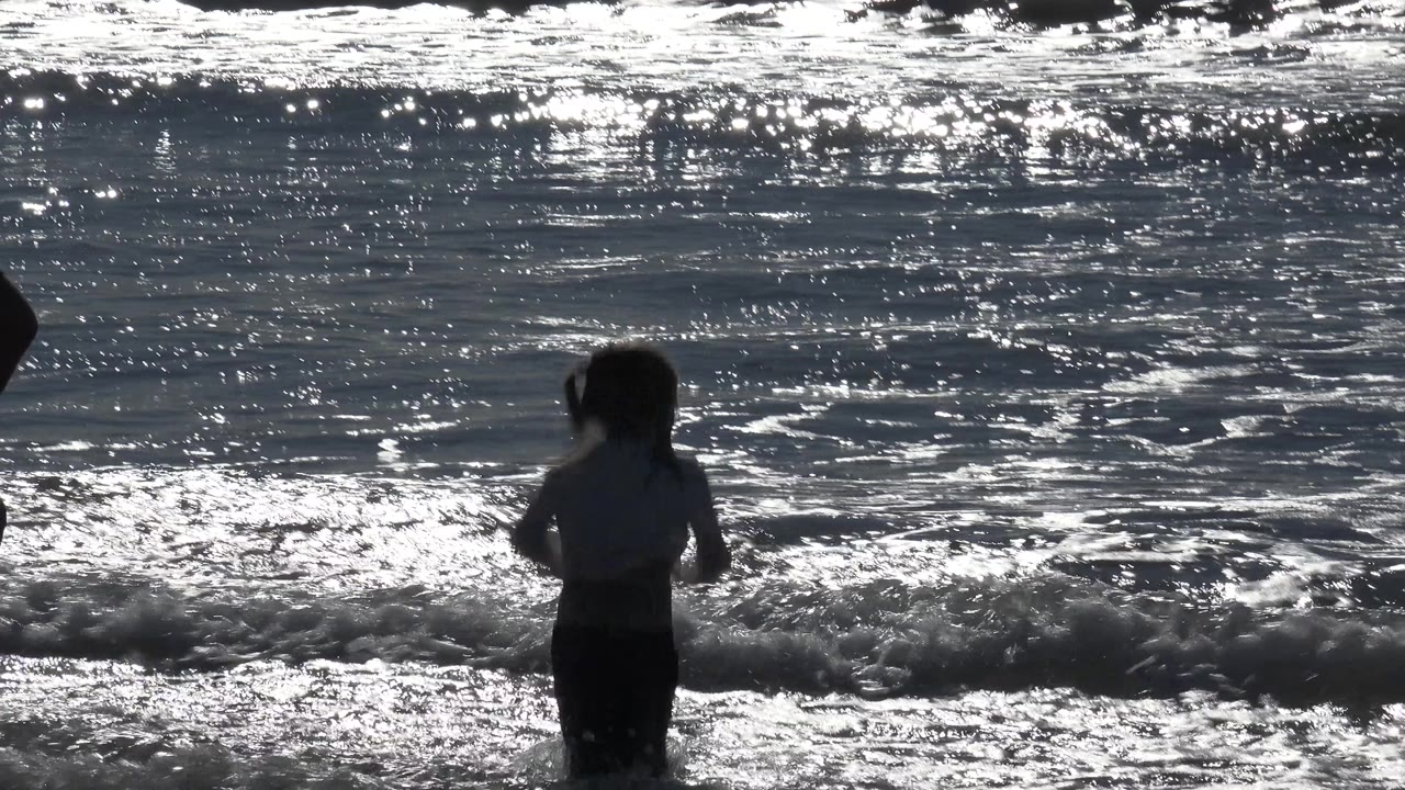 A small girl stands in shallow surf at Huntington Beach, California, with backlit water glittering behind her and a dark partial figure at the far left edge.