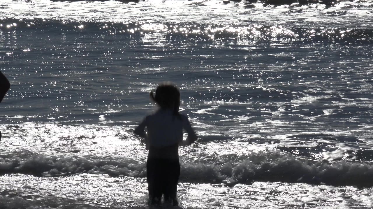 Small girl facing the waves as a foamy line breaks behind her.