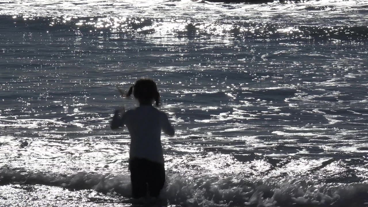 A small girl stands near the shore break at Huntington Beach, California, while a bright foamy crest crosses behind her and sun reflections fill the water.