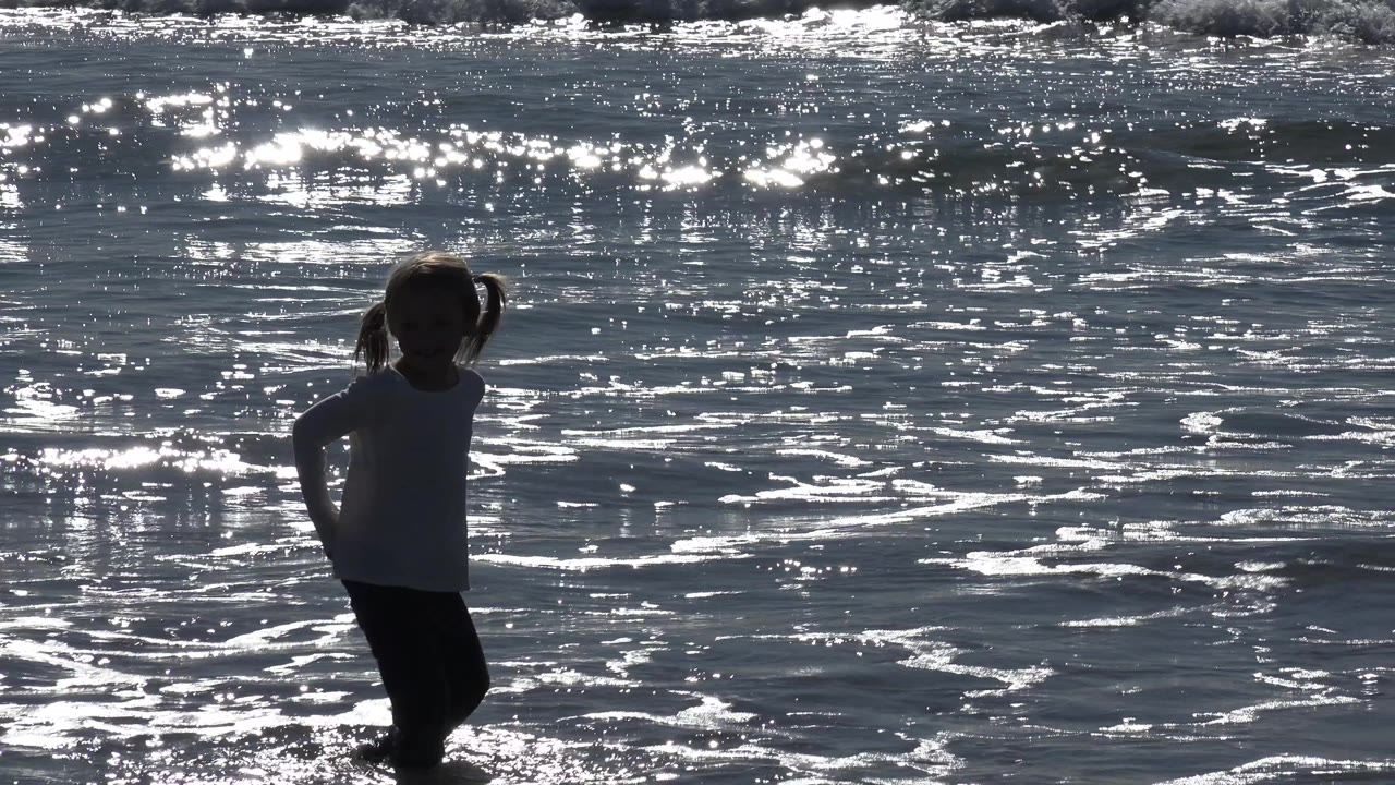 At Huntington Beach, California, a young girl in a white T-shirt stands near the left side of the frame in shallow surf, silhouetted against sparkling ocean water.