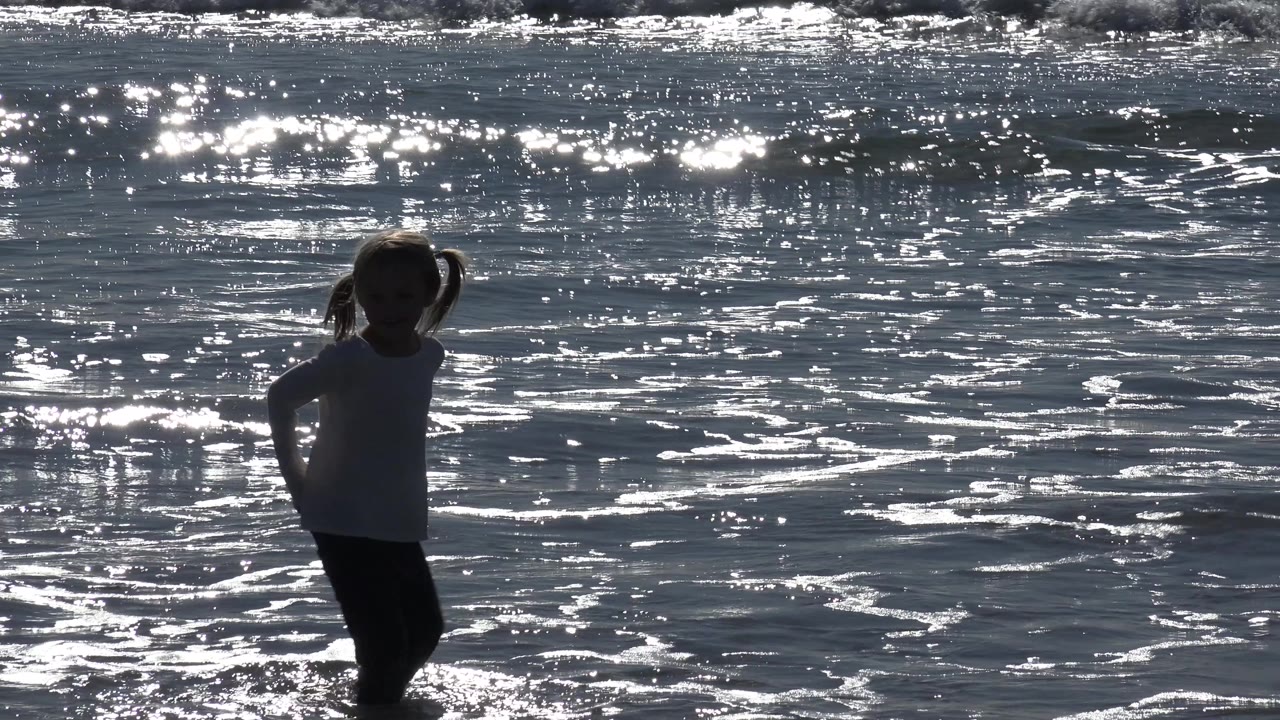 Young girl standing in shallow ocean water with sun glinting across the waves.