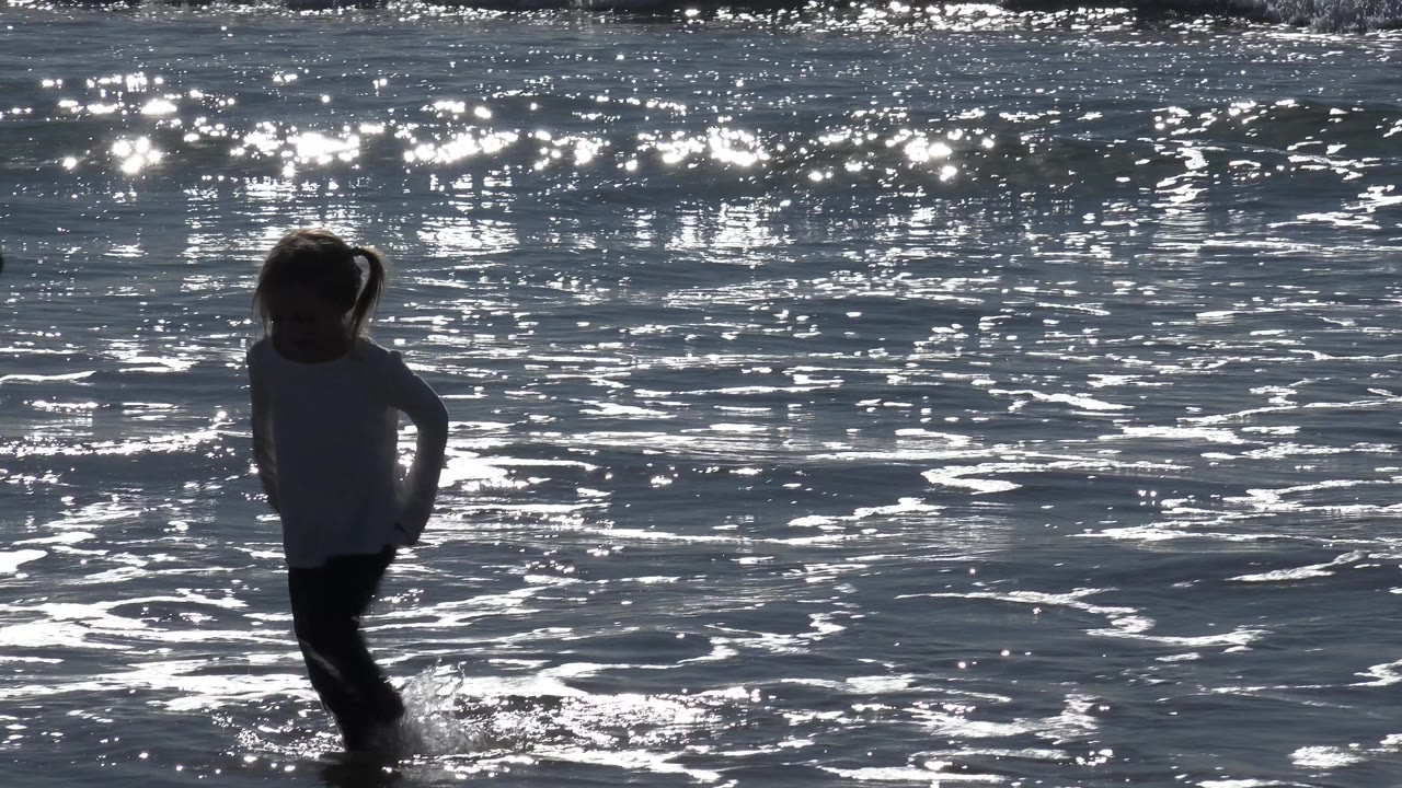 At Huntington Beach, California, the child shifts toward the right side of the frame, with small waves around her legs and bright reflections filling the background water.