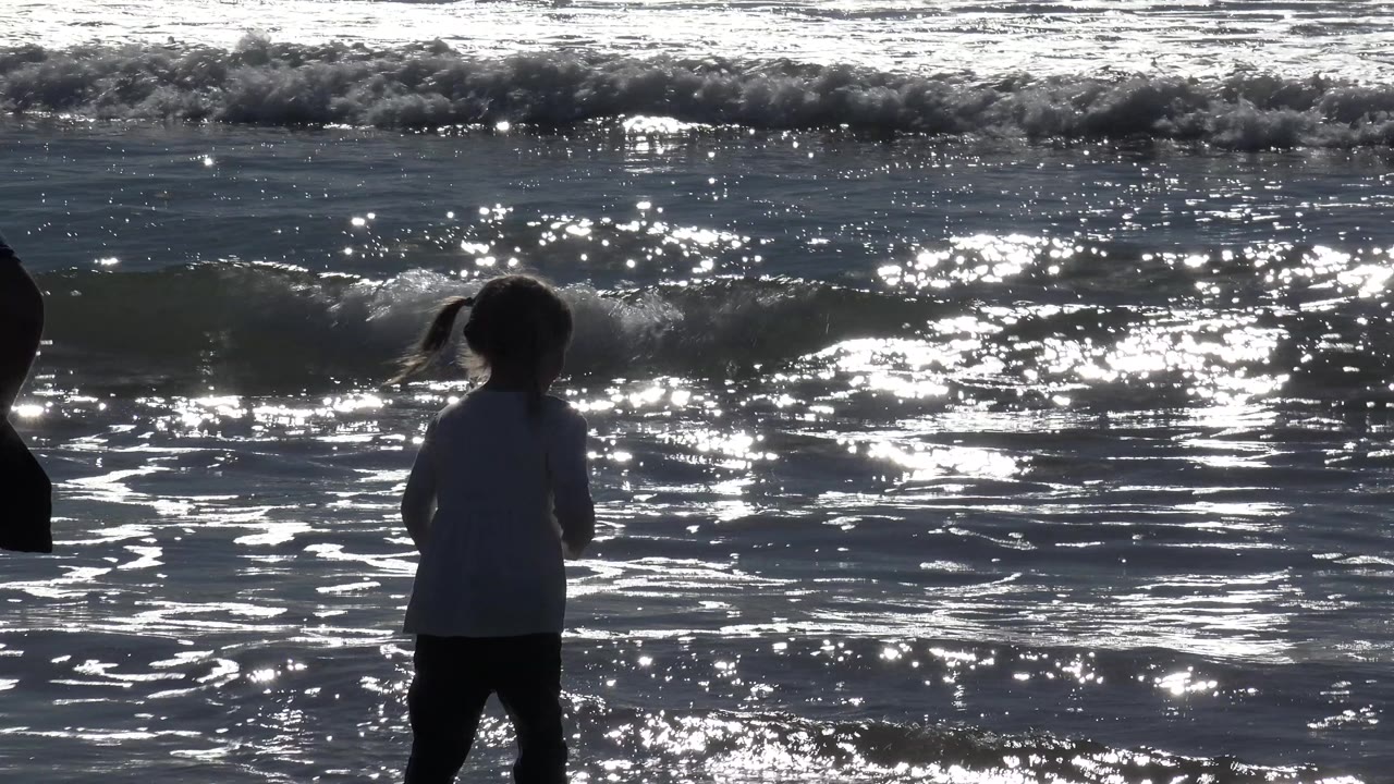 A young girl stands near the Huntington Beach shoreline in dark silhouette while sun glare sparkles across small breaking waves behind her.