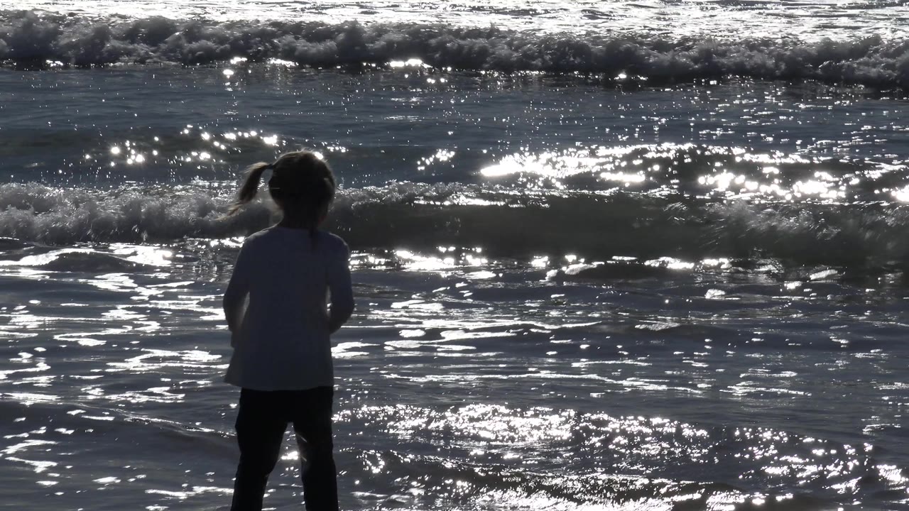 A young girl turns slightly at the Huntington Beach waterline, her pigtails visible in silhouette against bright reflected ocean light and foamy surf.
