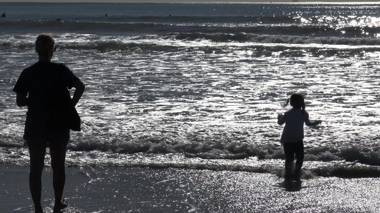 In Huntington Beach, California, a silhouetted adult stands at the left foreground while a child faces the incoming surf under strong reflected sunlight.
