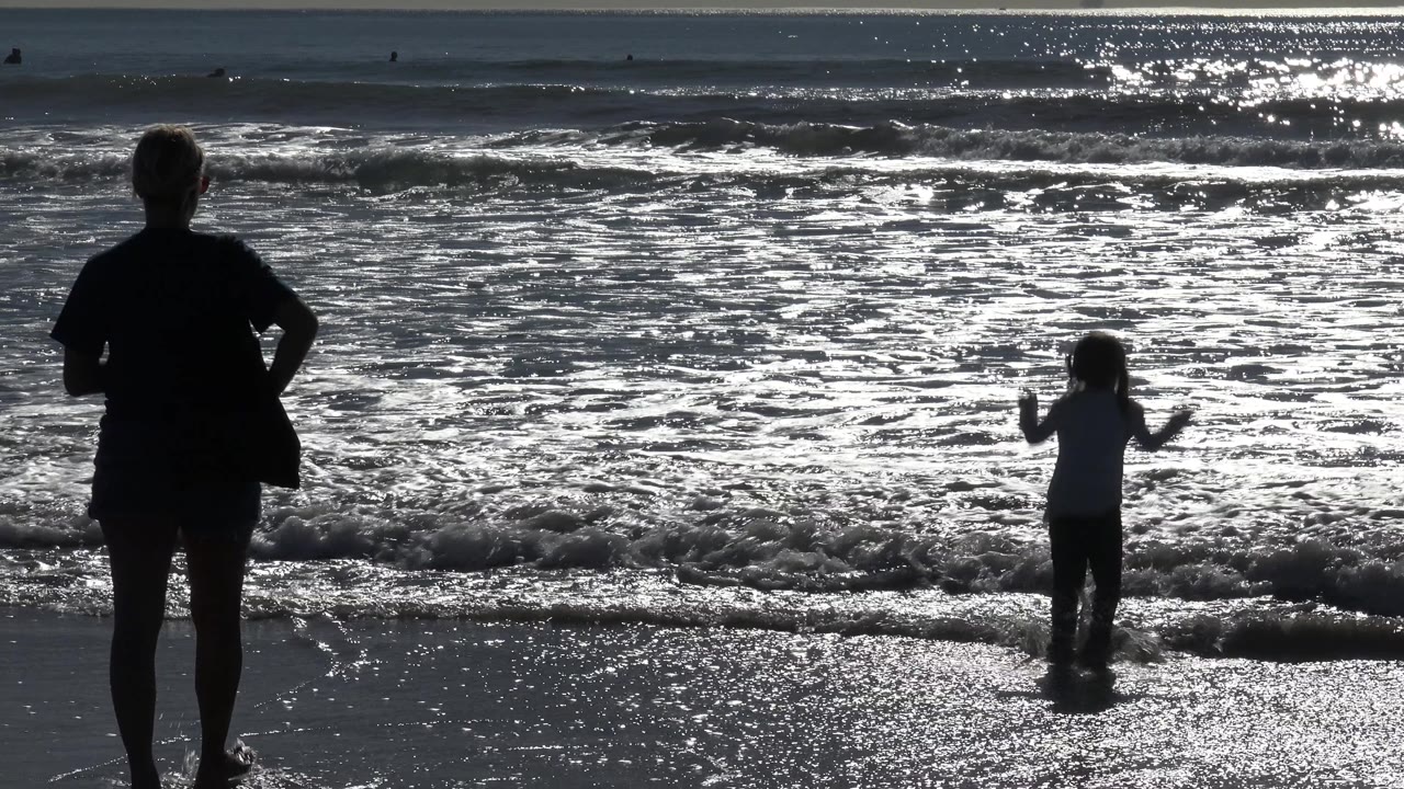 In Huntington Beach, California, the adult remains at left while the child stands closer to the center, framed by a small wave breaking behind them.