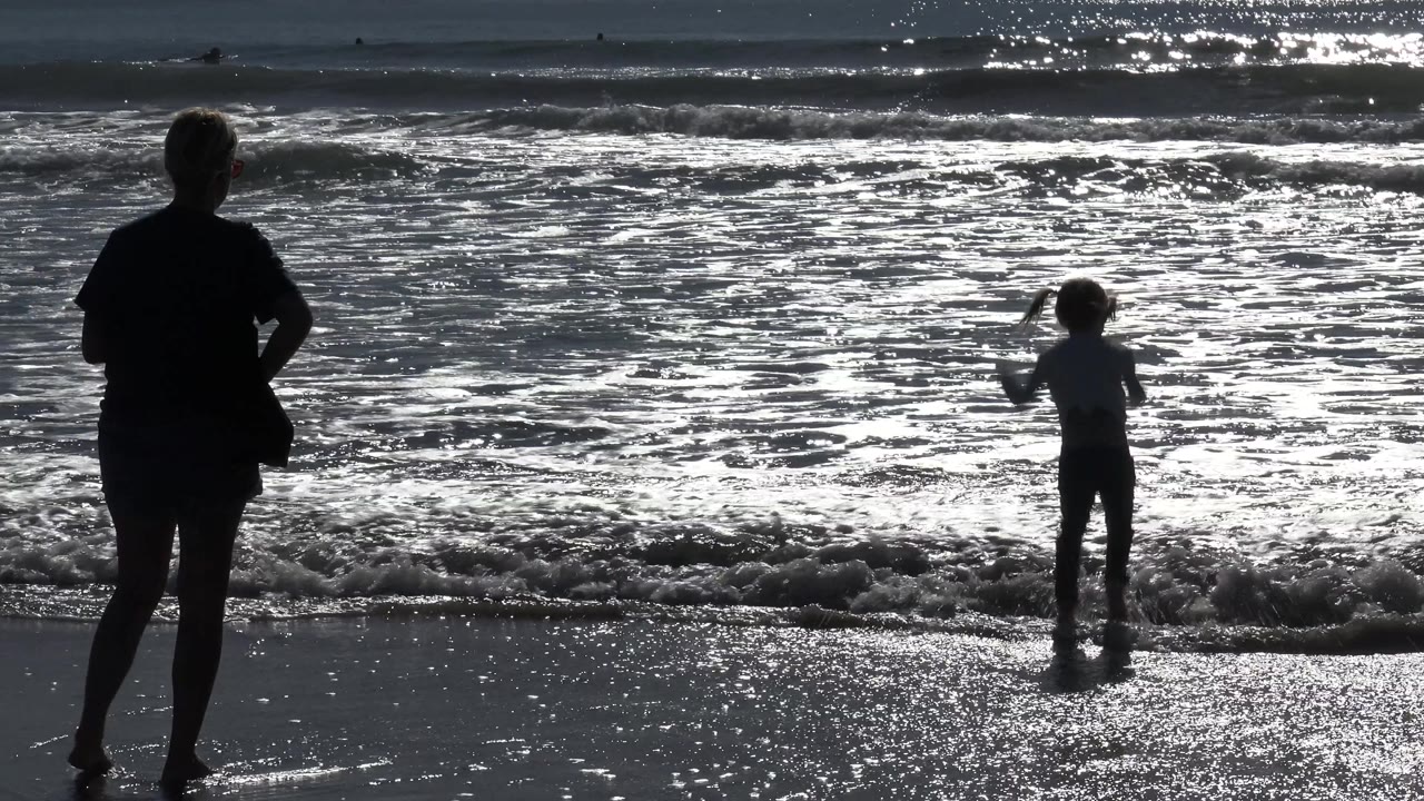 In Huntington Beach, California, the adult shifts slightly to the right foreground while the child raises an arm beside the foamy shorebreak.