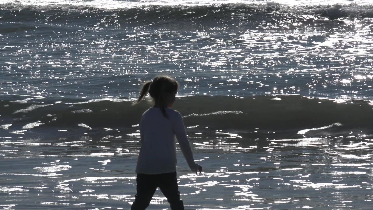 A backlit child stands near shallow surf at Huntington Beach, California, with bright ocean reflections and a low wave forming behind them.