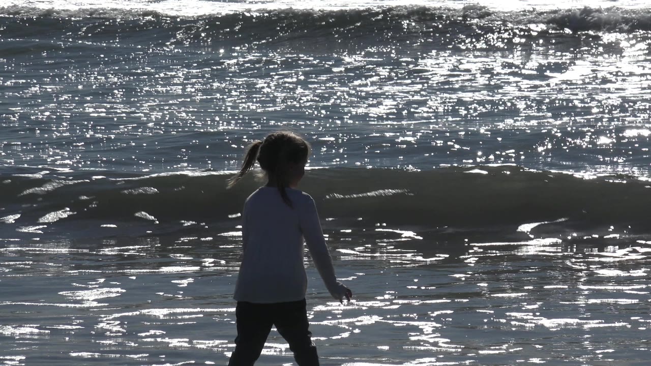 The child remains near the waterline at Huntington Beach, California, silhouetted against rippling silver water and a small shore break.