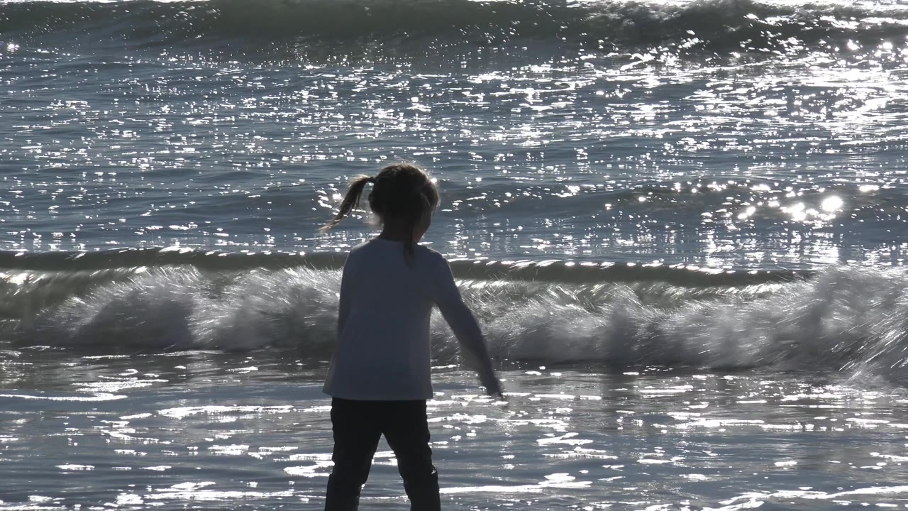 A child faces the surf at Huntington Beach, California, while a foamy wave curls across the background and sunlight flashes on the water.