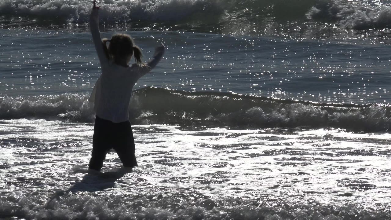 A child at Huntington Beach stands in bright shallow surf with both arms lifted as sunlight glitters across the ocean behind her.