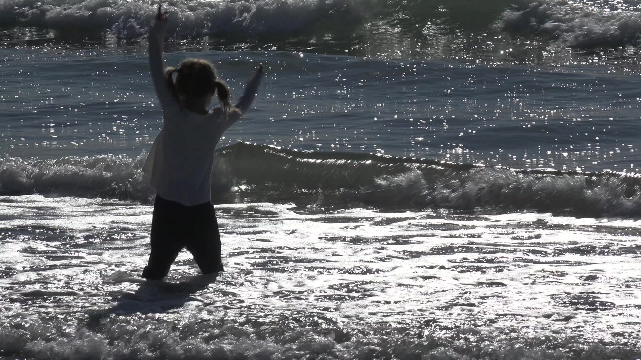 A child at Huntington Beach remains near the foamy edge of the water with raised arms while a low wave breaks across the background.
