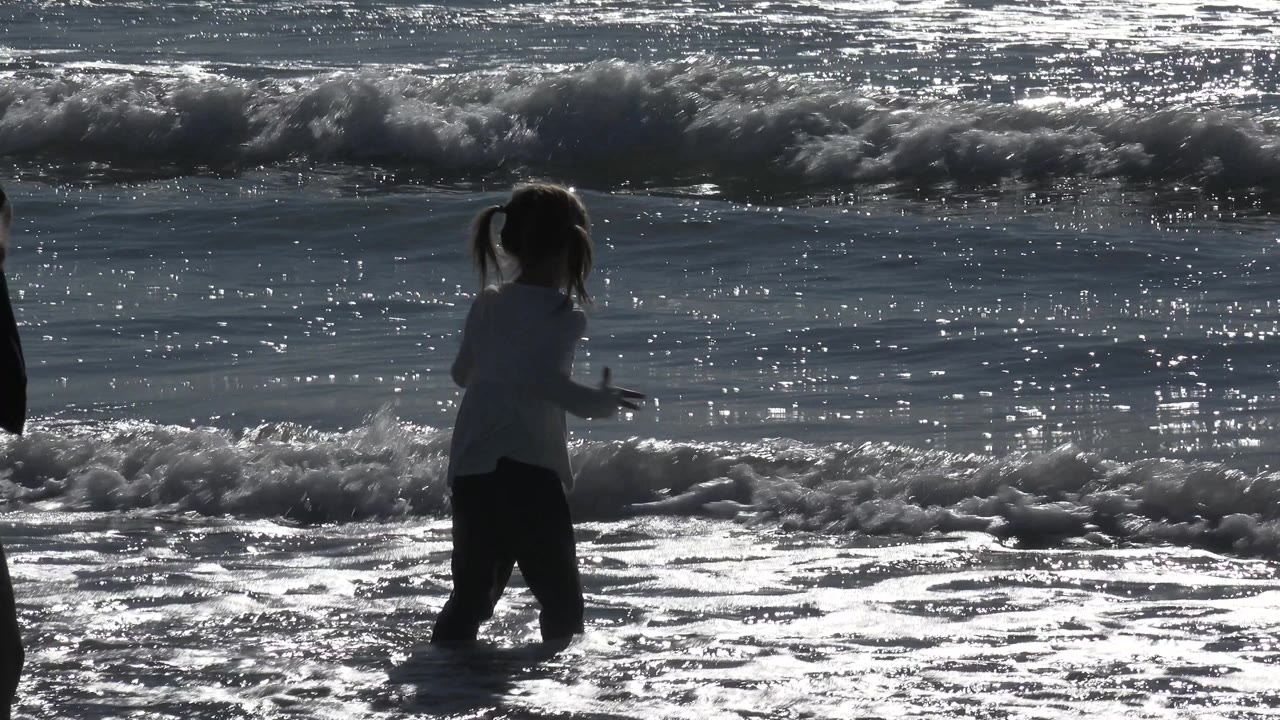 A child at Huntington Beach turns sideways in ankle-deep surf, with a bright band of foam crossing the middle of the frame.