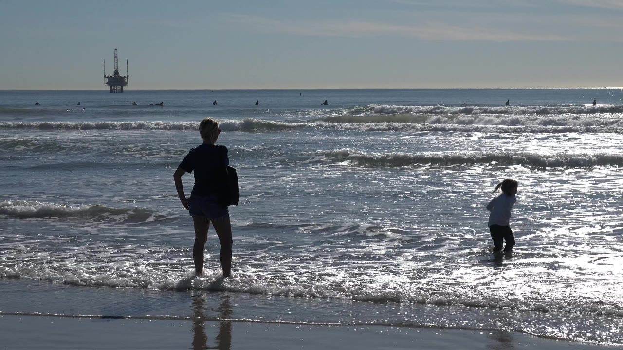 At Huntington Beach, an adult silhouette stands at the waterline while a child wades in bright reflective surf, with surfers and an offshore platform in the distance.