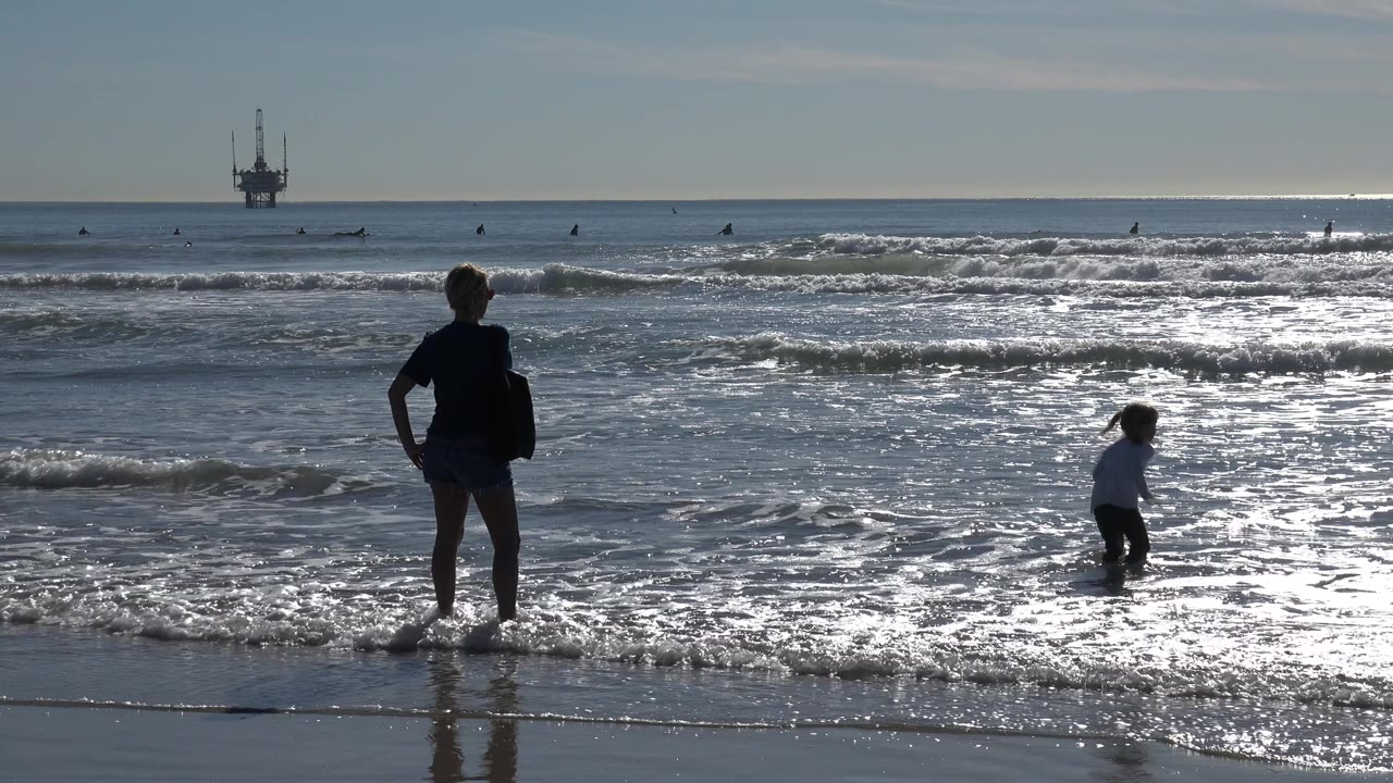 At Huntington Beach, the child moves through the foamy shallows as the adult remains near the left foreground and the offshore platform sits beyond the breaking waves.