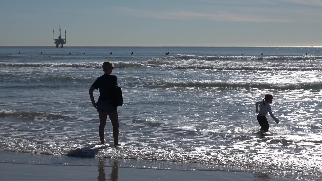 At Huntington Beach, the adult stands with hands near the hips as the child appears farther right in the shallow surf, with a bright band of reflected light across the ocean.