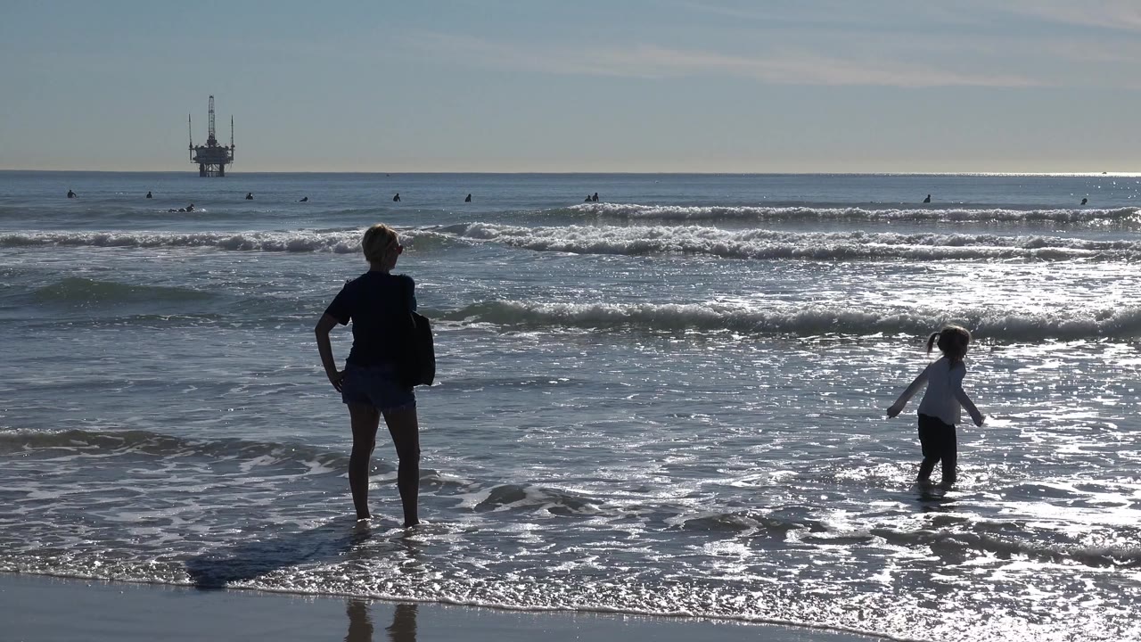 Child wades in foamy water as an adult stands at the left edge of the surf.