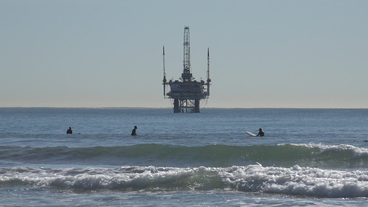 Surfers sit in the water off Huntington Beach, California as a low wave rises in front of them and an offshore platform stands on the horizon.