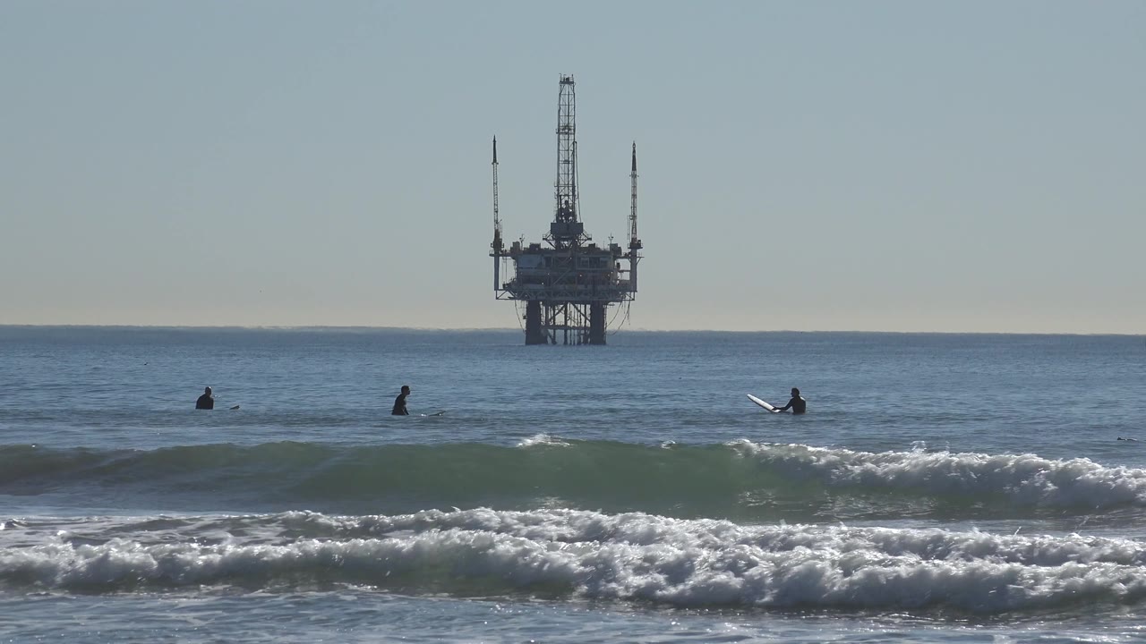 A white surfboard stands out on the right side of the Huntington Beach lineup as a curling wave and offshore platform define the scene.