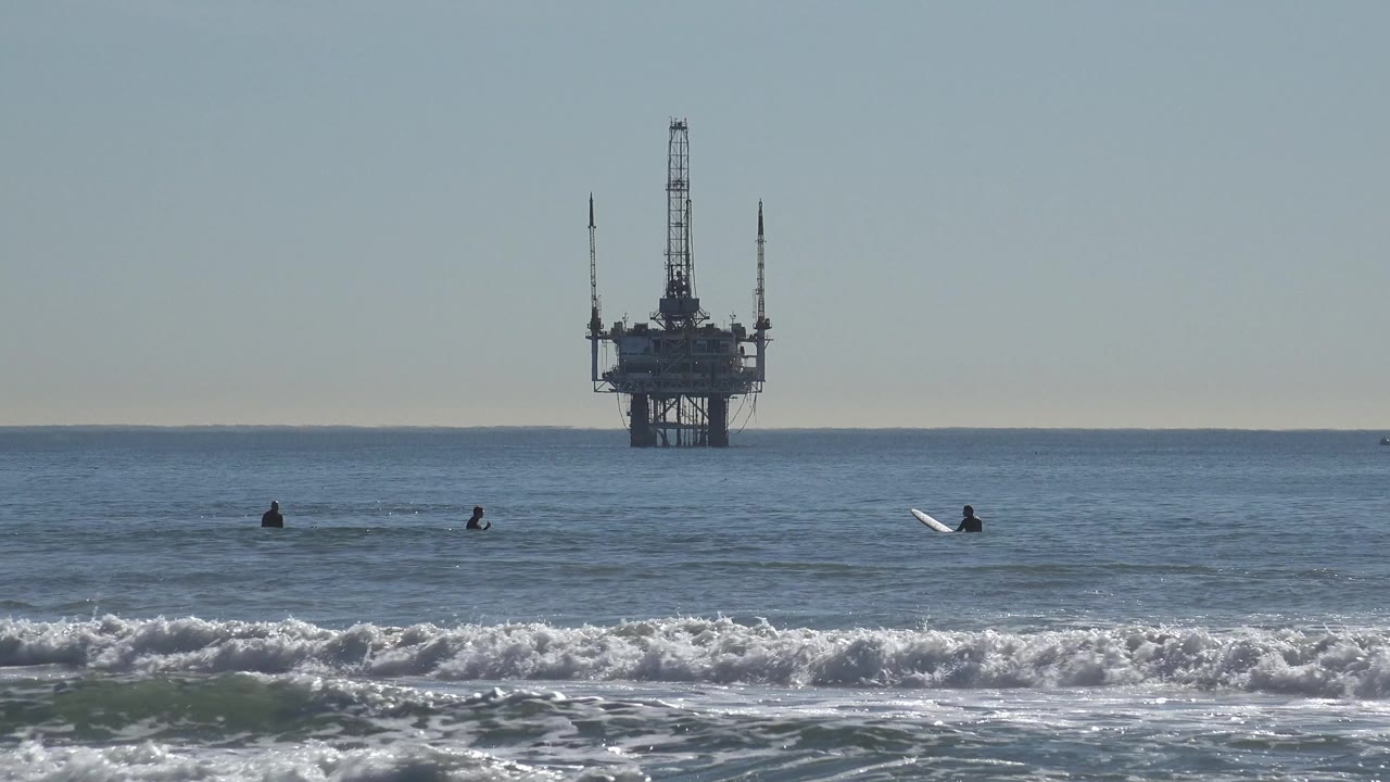 Huntington Beach ocean view with several small surfers in the water, white shore break in front, and an offshore oil rig centered near the horizon.