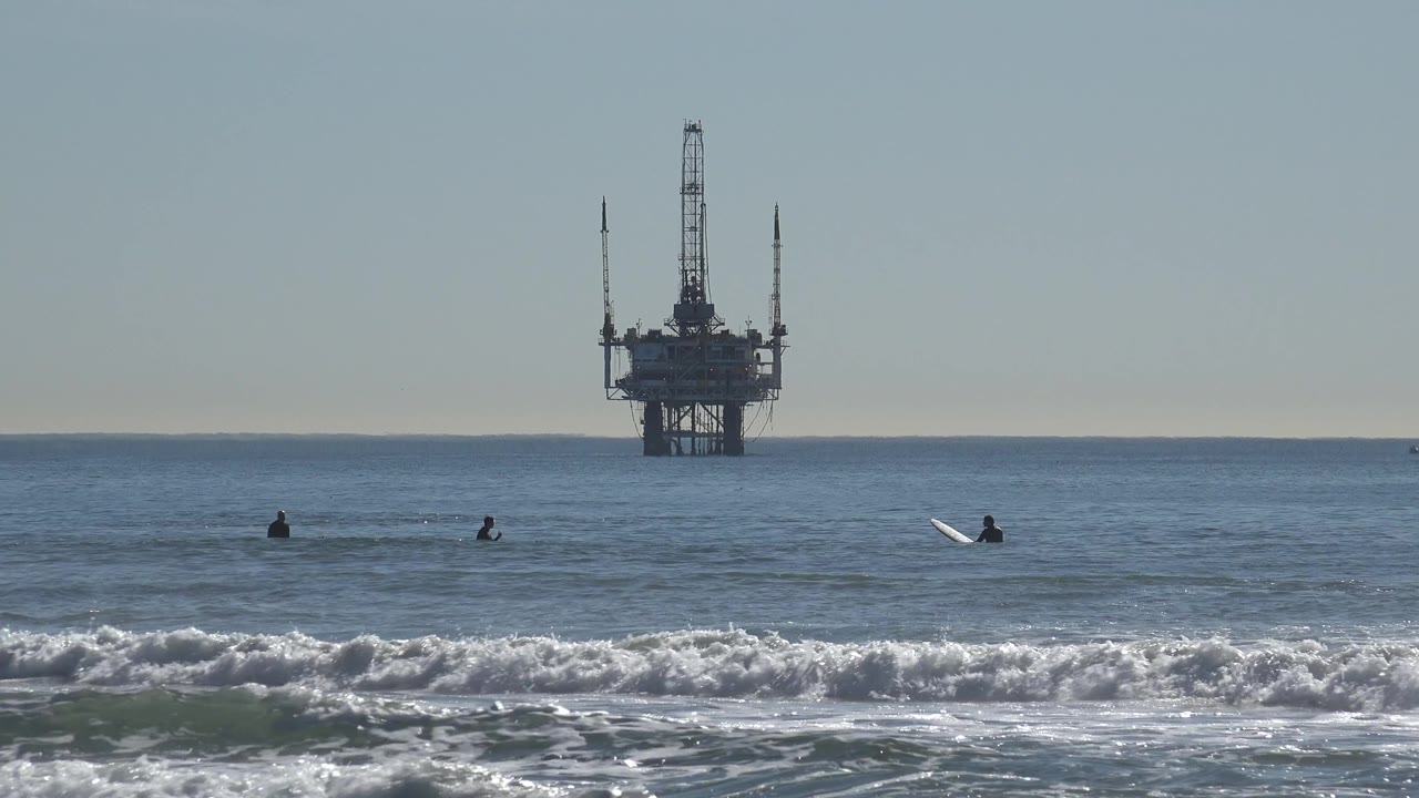 Huntington Beach surf line with silhouetted surfers spaced across the water and a dark offshore oil rig rising behind them under pale blue sky.