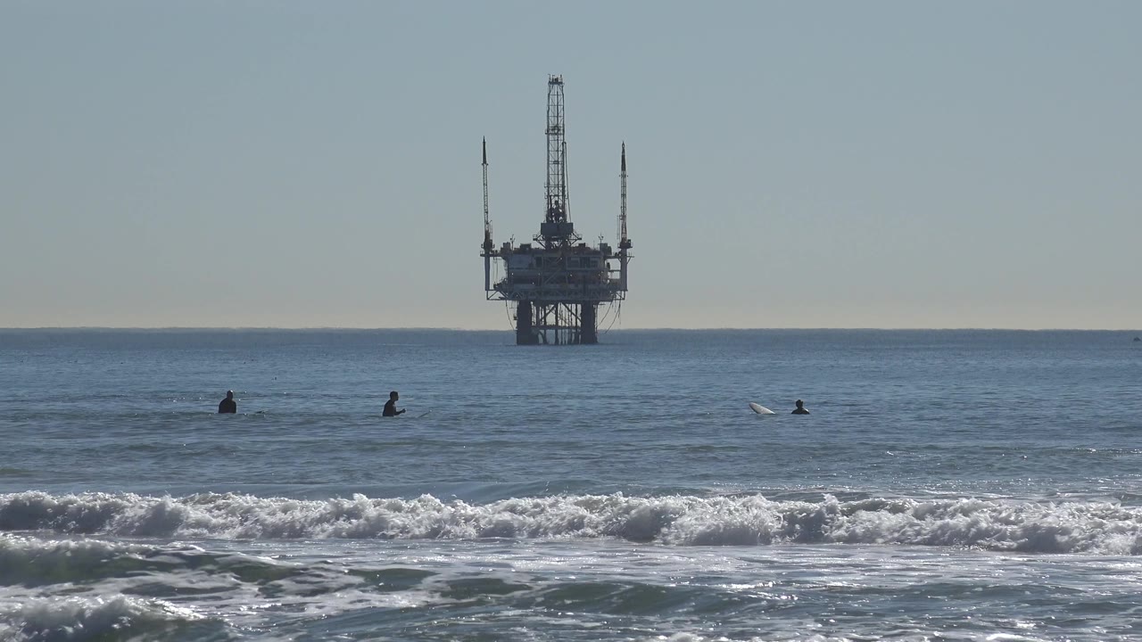Huntington Beach water scene with a low swell crossing the foreground, scattered surfers in the midground, and the offshore oil rig shifted toward the right side of frame.
