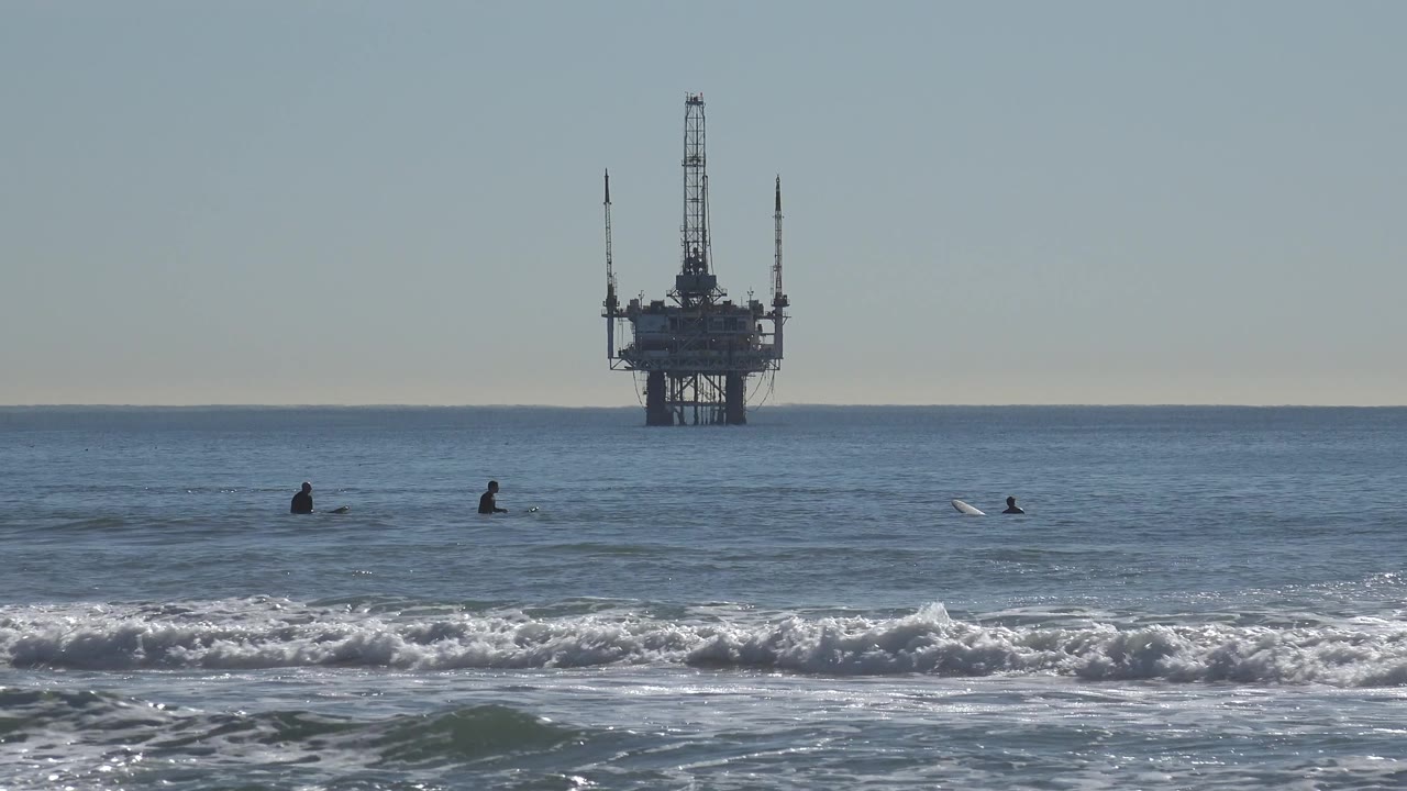 Offshore oil rig behind quiet water and surfers near the break.