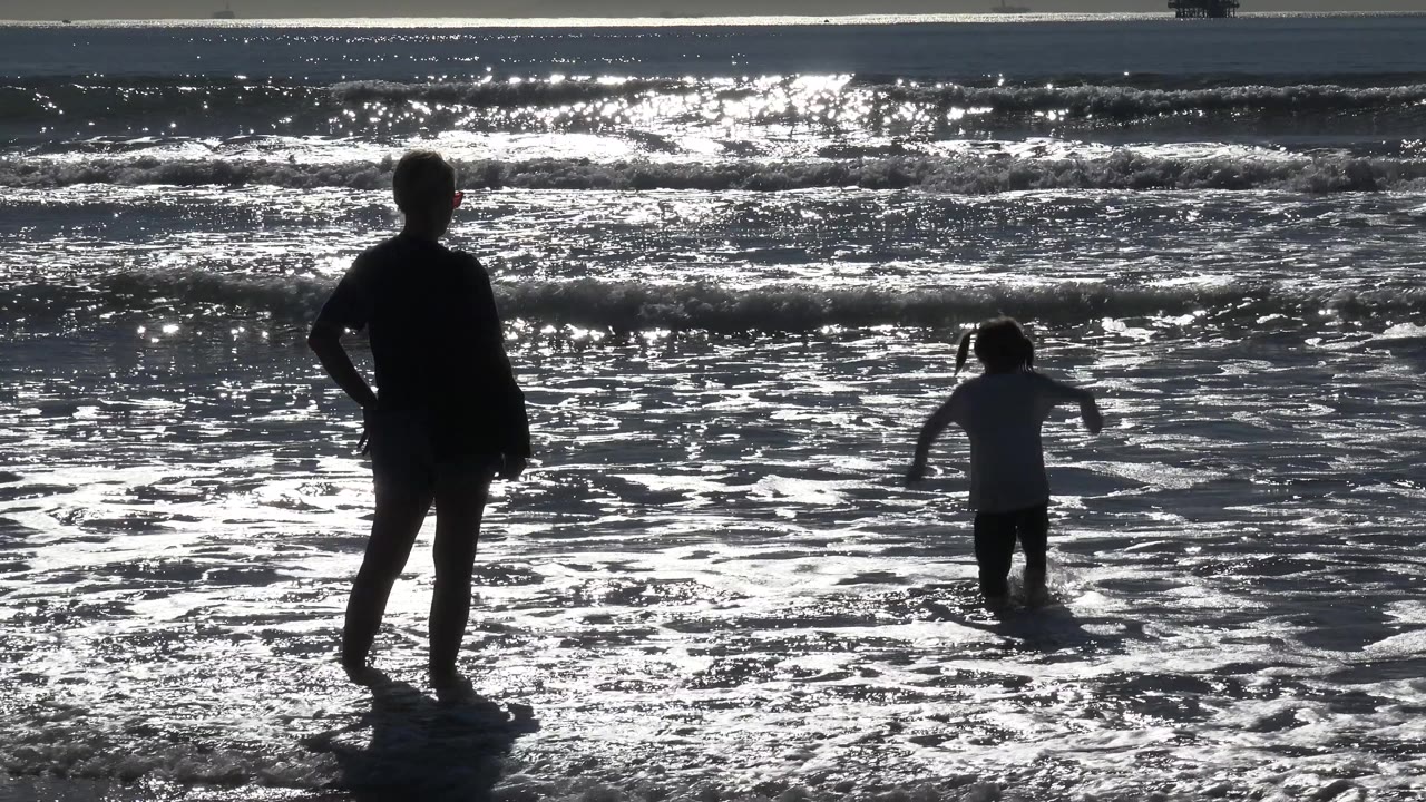 A silhouetted adult stands at left while a young girl faces the waves in shallow surf at Huntington Beach, with strong glare on the wet sand.