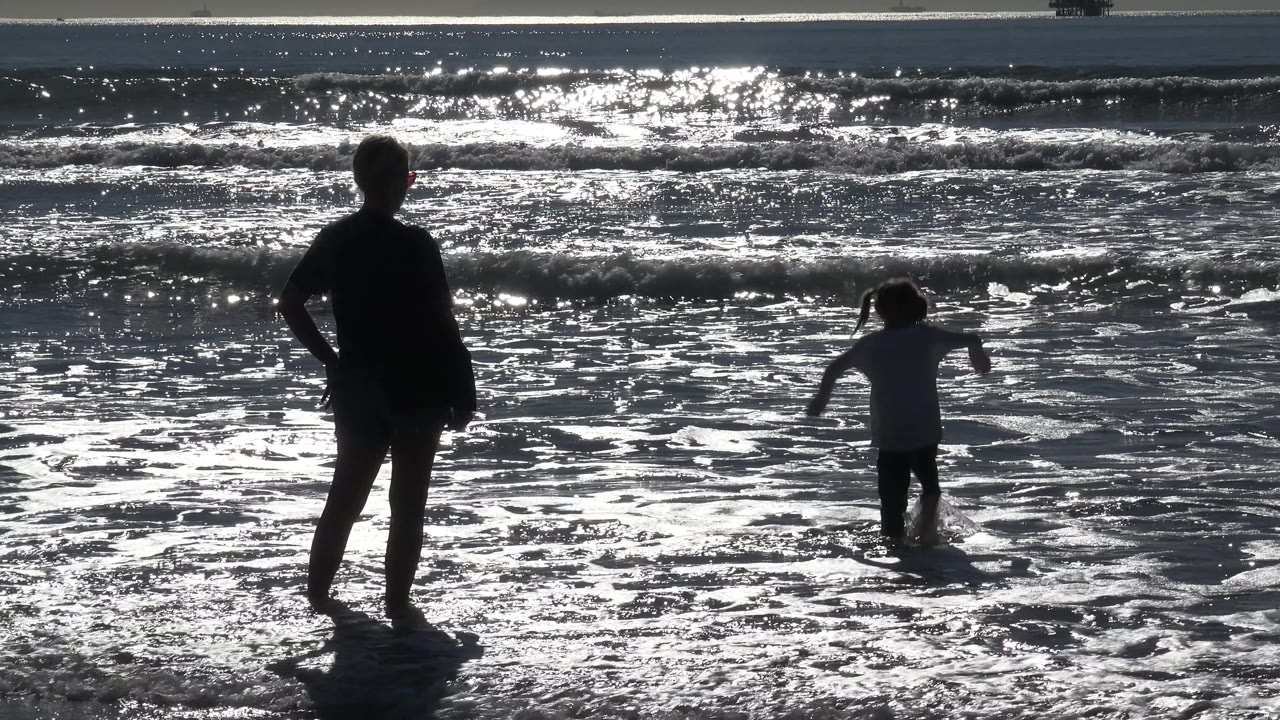 The young girl raises her arms in the shallow water as the adult remains silhouetted at left at Huntington Beach, with small waves breaking behind her.