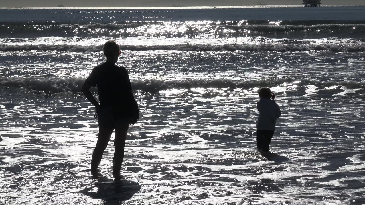 An adult silhouette holds position near the left foreground while the young girl turns to the side in the Huntington Beach wash, with bright wave highlights across the frame.