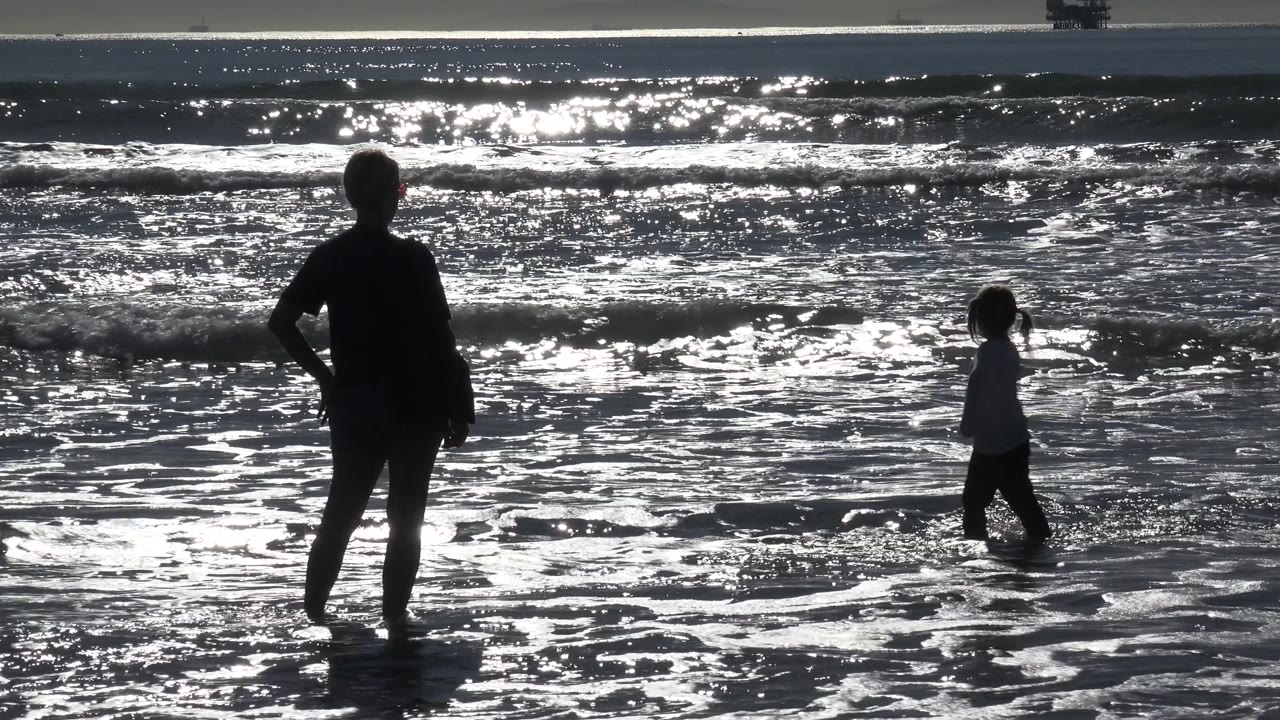 Young girl walks through shallow water with a silhouetted adult in the foreground.