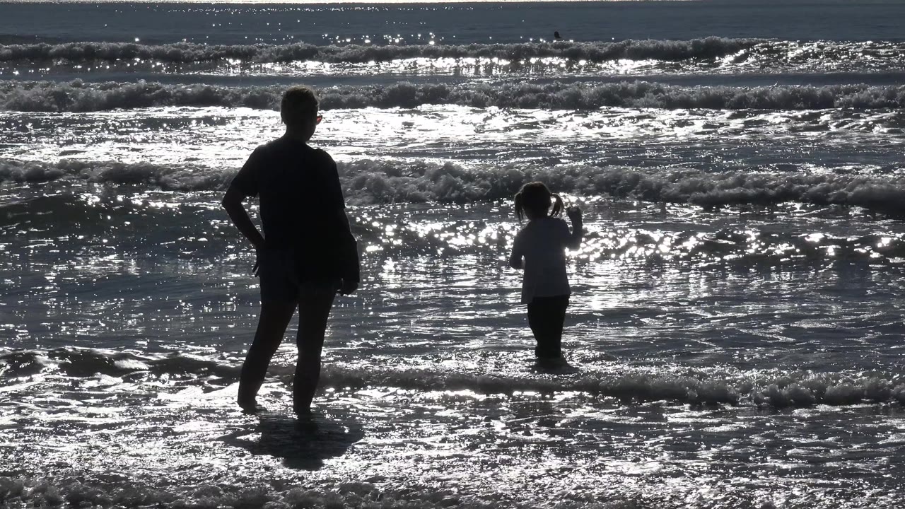 At Huntington Beach, California, an adult stands at left while a child faces the breaking waves in bright reflected surf.