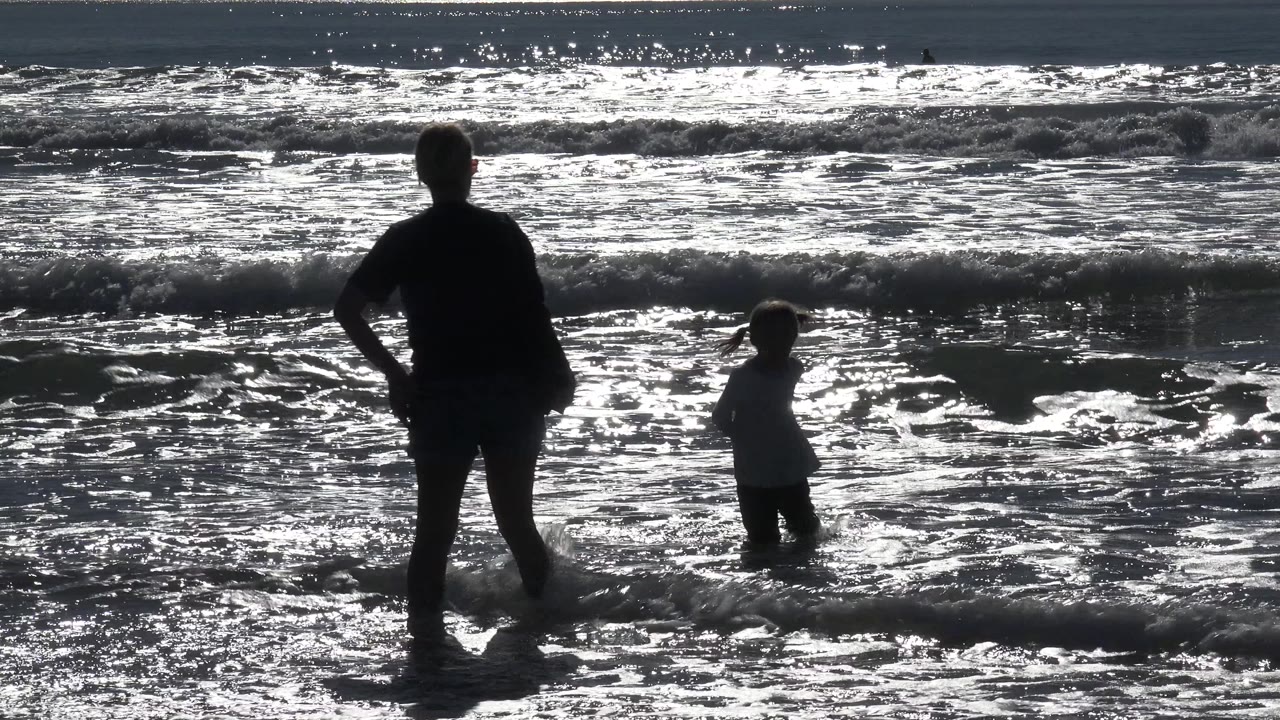 A silhouetted woman and child stand in shallow waves at Huntington Beach, California, with bright reflected sunlight covering the ocean behind them.